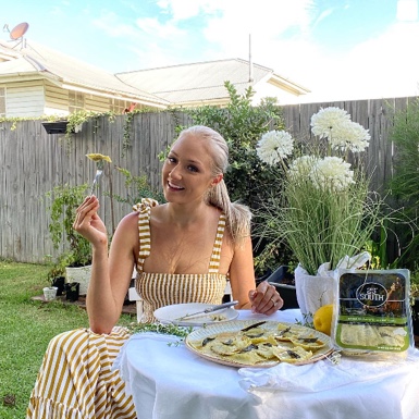 A woman in a yellow and white striped dress sits at an outdoor table, smiling and holding a fork with food. - Link to Instagram post