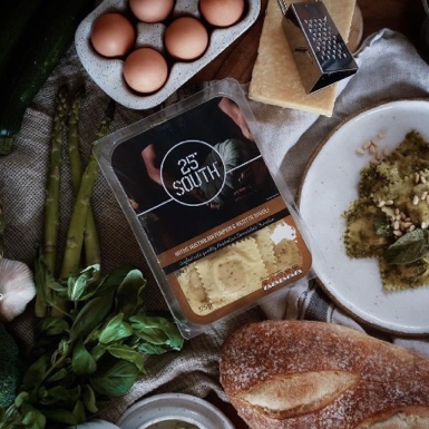A package of 25° South ravioli surrounded by eggs, asparagus, zucchini, basil, garlic, a loaf of bread, cheese, a grater, and a plate of pasta. - Link to Instagram post