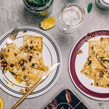 Two plates of ravioli with sauce and nuts are set on a table with a glass of water, lemon slices, cutlery, and a jar of greens nearby. - Link to Instagram post