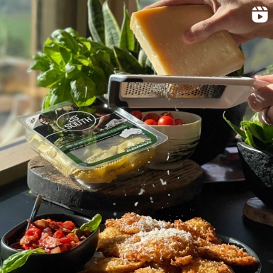 A hand grates cheese over a plate of breaded food, surrounded by fresh basil, packaged pasta, cherry tomatoes, and a bowl of chopped vegetables. - Link to Instagram post