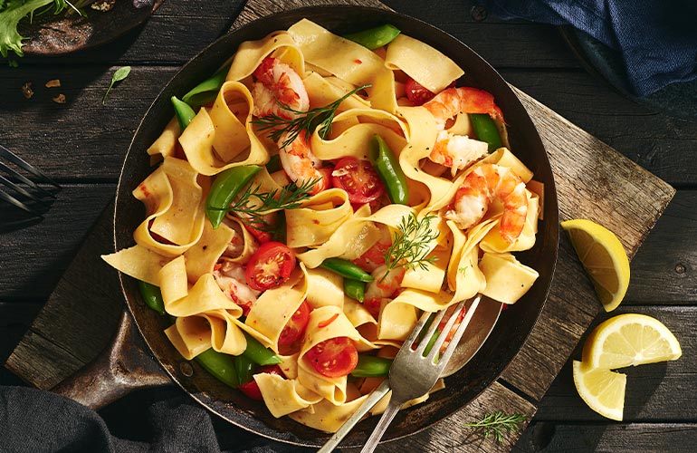 A skillet of pappardelle pasta with shrimp, cherry tomatoes, and snap peas, garnished with fresh dill, sits on a wooden board next to lemon wedges and cutlery.
