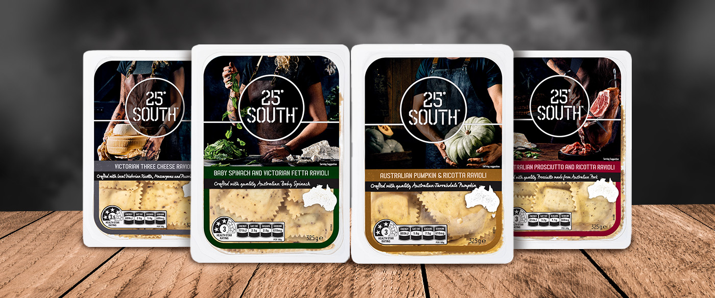 Four packages of 25° South branded ravioli in different flavors are displayed upright on a wooden surface with a dark, blurred background.