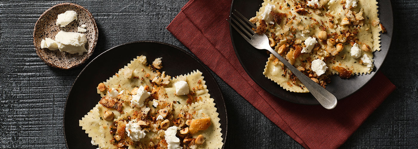 Two plates of Prosciutto and Ricotta Ravioli with Hazelnut Pangrattato next to a bowl of hazelnuts and a plate of proscuitto on a black surface.